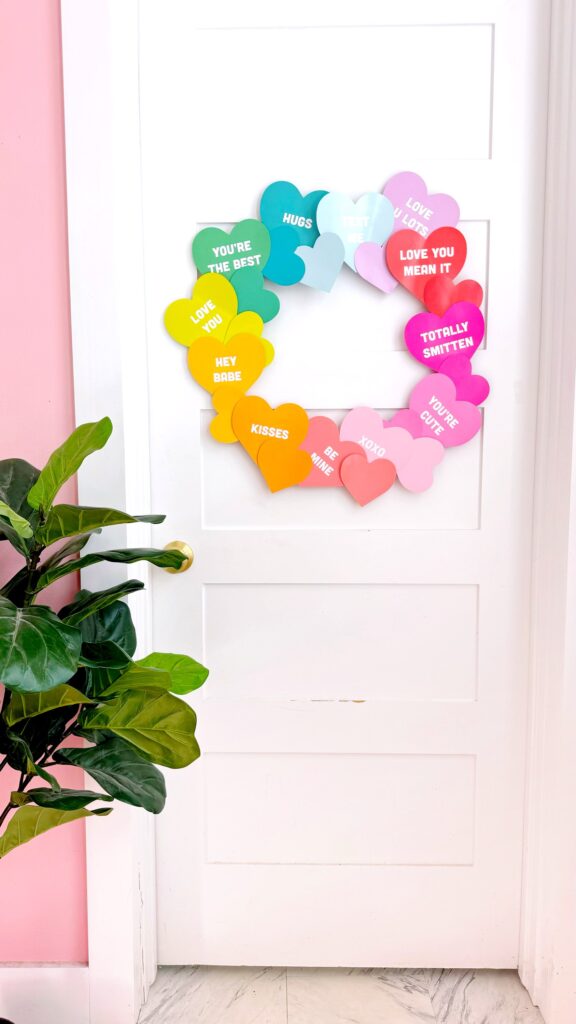 picture of white door with a wreath made of rainbow paper hearts with cute love related phrases